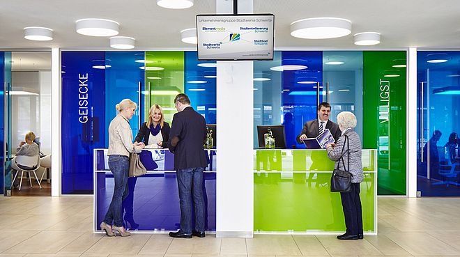 Kundenschalter mit blauen und grünen Glaswänden, Beschriftungen 'GEISECKE' und 'WILLIGST', Personen an den Schaltern im Gespräch