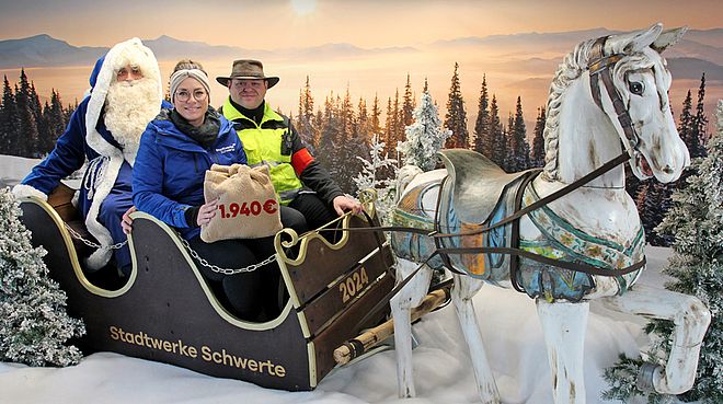 Drei Personen sitzen in einem Schlitten mit der Aufschrift 'Stadtwerke Schwerte', der von einem weißen Pferd gezogen wird, vor einer Winterlandschaft mit verschneiten Bäumen und Bergen im Hintergrund.