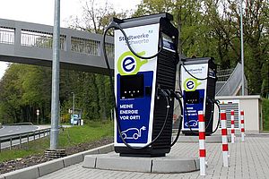 Zwei Ladesäulen der Stadtwerke Schwerte mit der Information "Mehr Energie vor Ort". Die Ladesäulen und dazugehörigen Parkplätze befinden sich neben einer Schnellstraße und einer Fußgängerbrücke